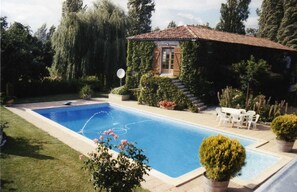 Ein rechteckiger Pool mit Fliesen umgeben, mit einer gepflegten Rasenfläche und verschiedenen Pflanzen. Ein kleines Gebäude mit einem gefliesen Dach und Holzläden steht in der Nähe, sowie ein Tisch mit Stühlen.