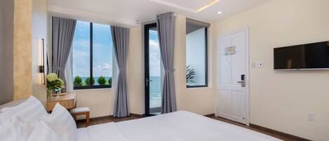 A modern hotel room with a large bed, a desk, a chair, a TV, and a view of the sea.