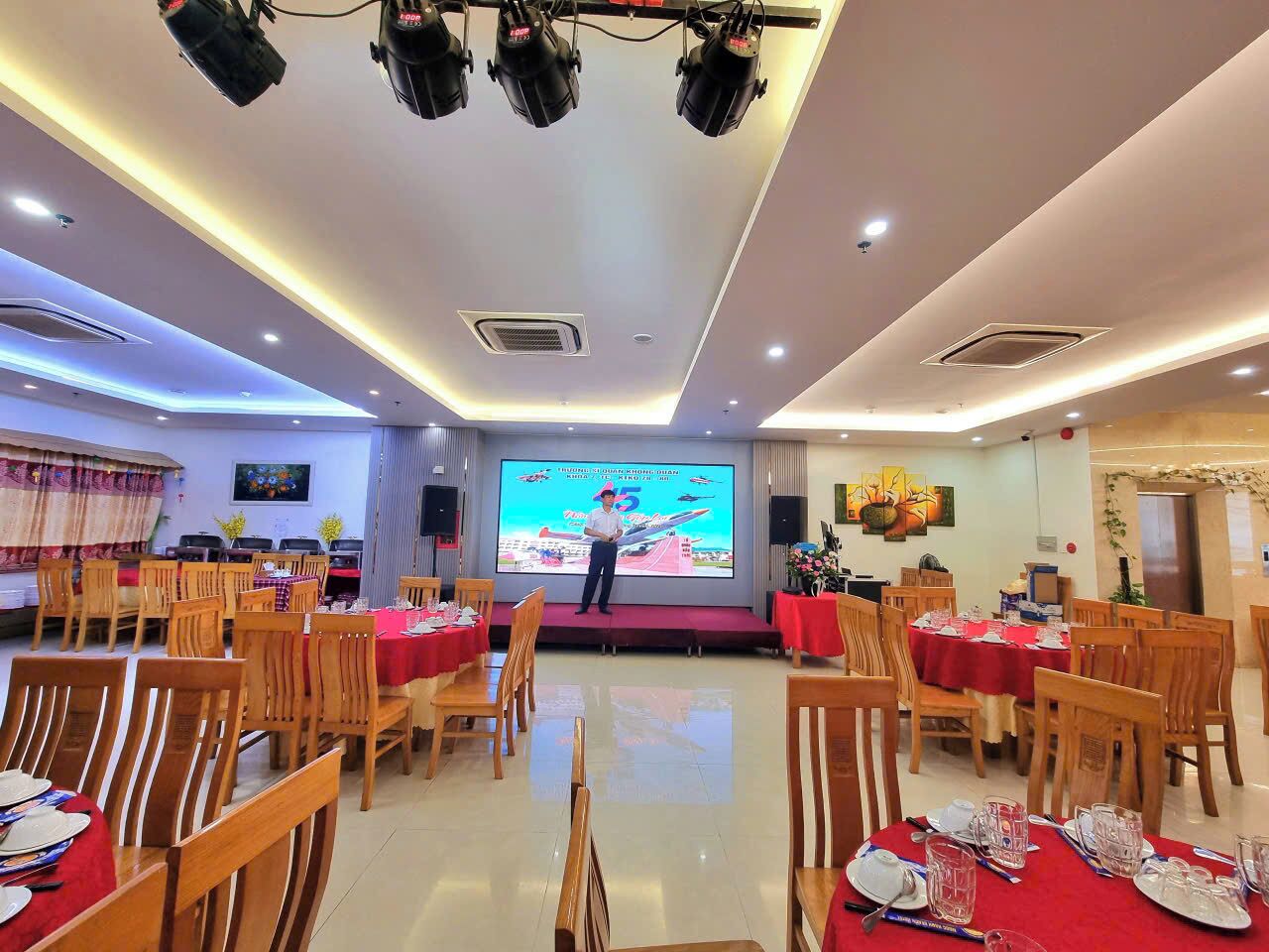 A conference room with a stage, multiple tables with red tablecloths, wooden chairs, and a large screen displaying a presentation.