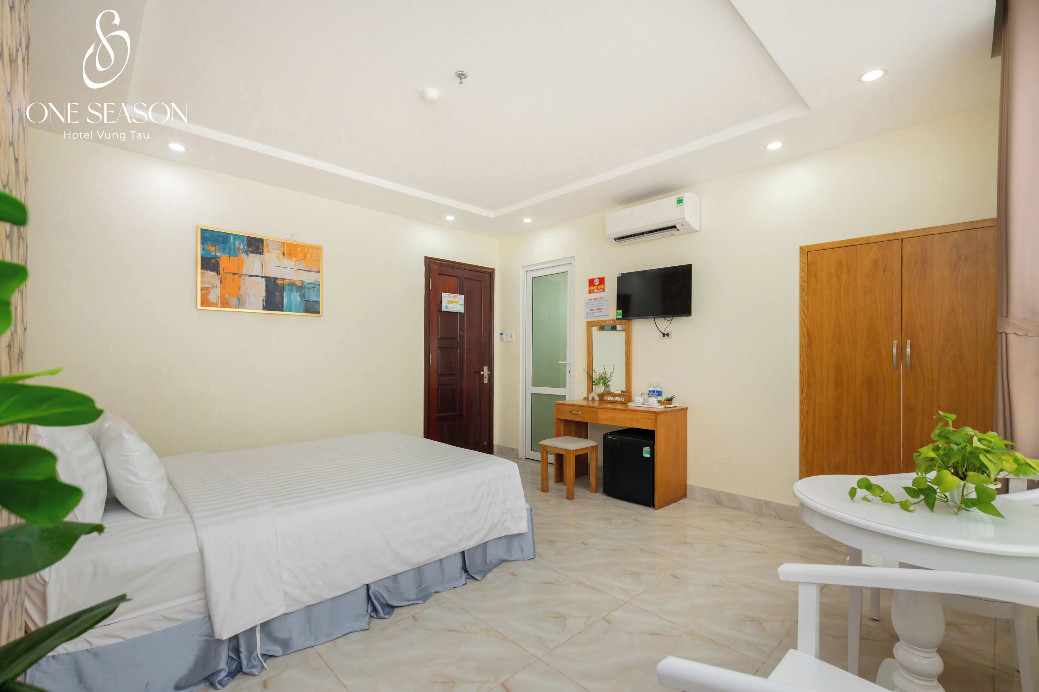 A hotel room with a bed, desk, chair, TV, wardrobe, and a small table with a plant.