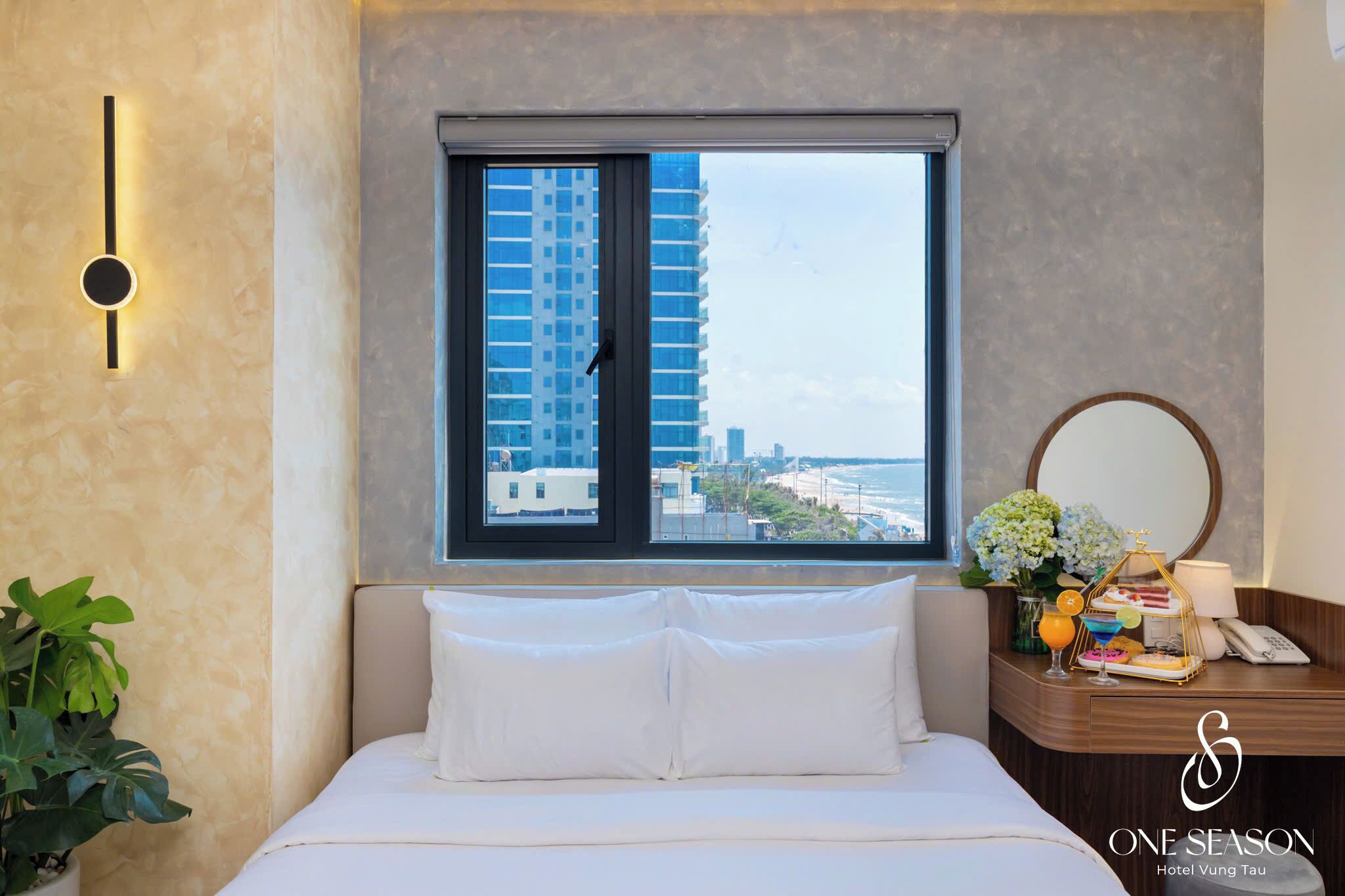 A hotel room with a bed, a bedside table with a lamp, a mirror, and a window with a city view.