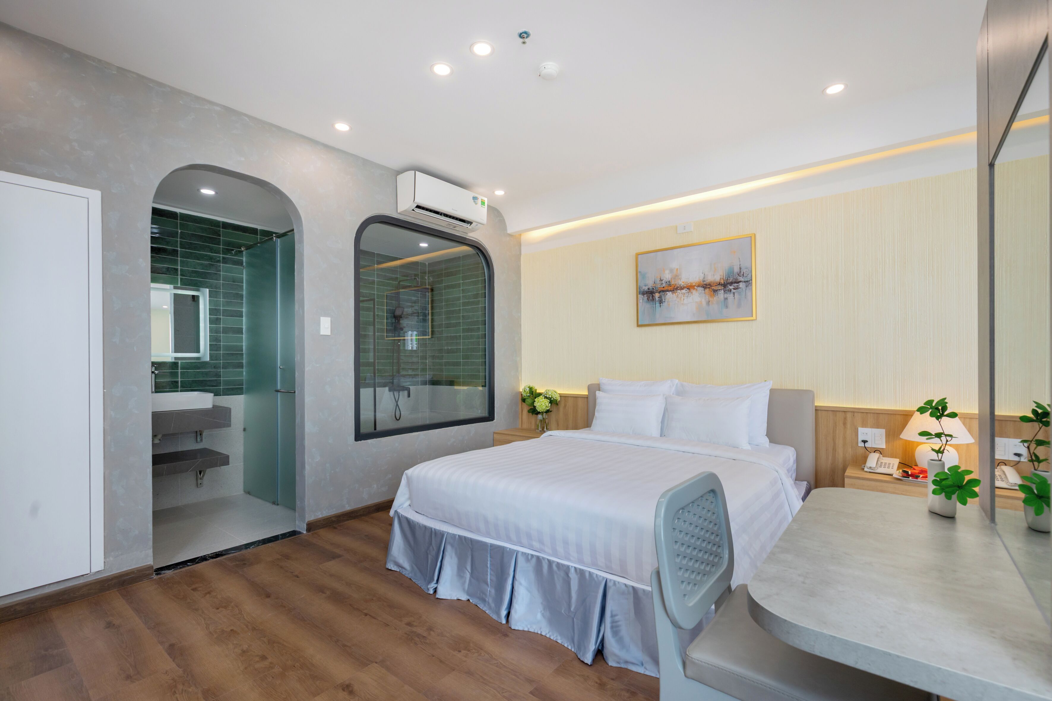 Ein modernes Hotelzimmer mit einem großen Bett, einem Schminktisch mit Spiegel, einer Duschkabine und einem kleinen Schreibtisch mit Lampe und Pflanzen.