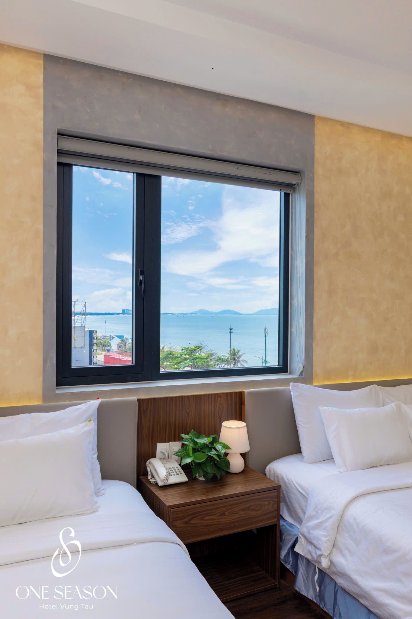 A hotel room with two beds, a wooden nightstand with a lamp and phone, and a window with a view of the sea and distant land.