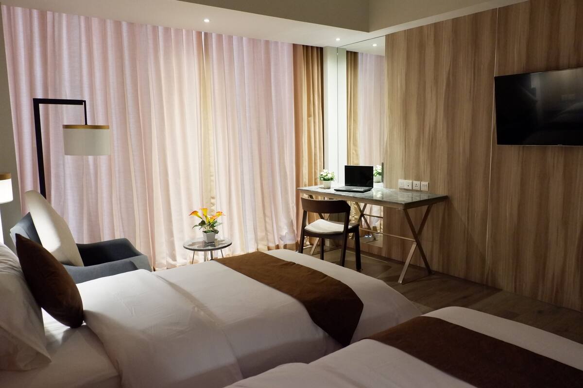 Een hotelkamer met twee bedden, een bureau met een laptop, een televisie en een klein tafeltje met bloemen.