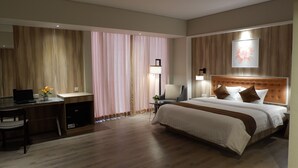 Une chambre d’hôtel avec un grand lit, un bureau avec une chaise, une petite table avec une lampe et une fenêtre avec des rideaux.