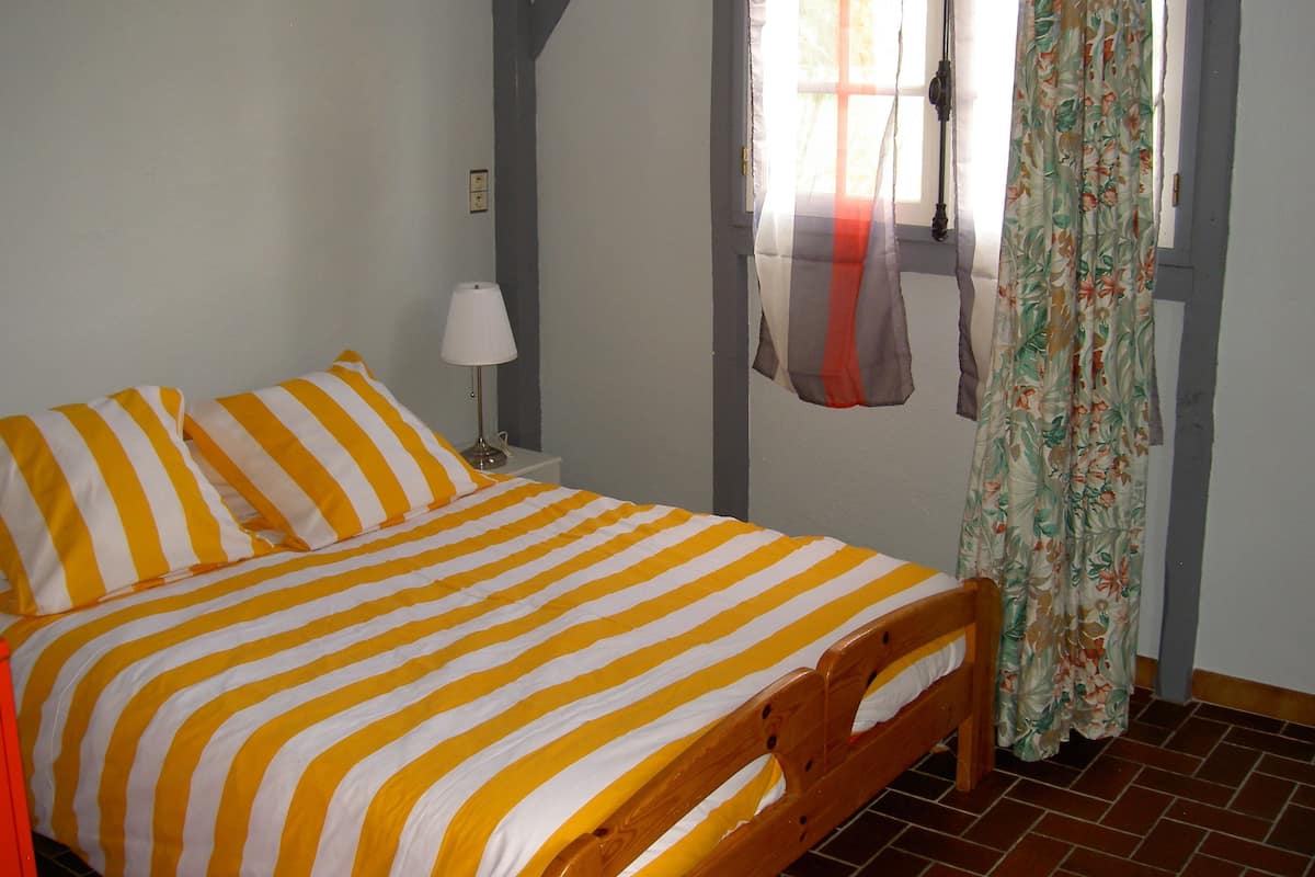 Ein Zimmer mit einem Bett, ausgestattet mit gelb-weiß gestreifter Bettwäsche, einem Holz-Kopfteil, einem Nachttisch mit Lampe, einem Fenster mit floralen Vorhängen und einem gefliesten Boden.