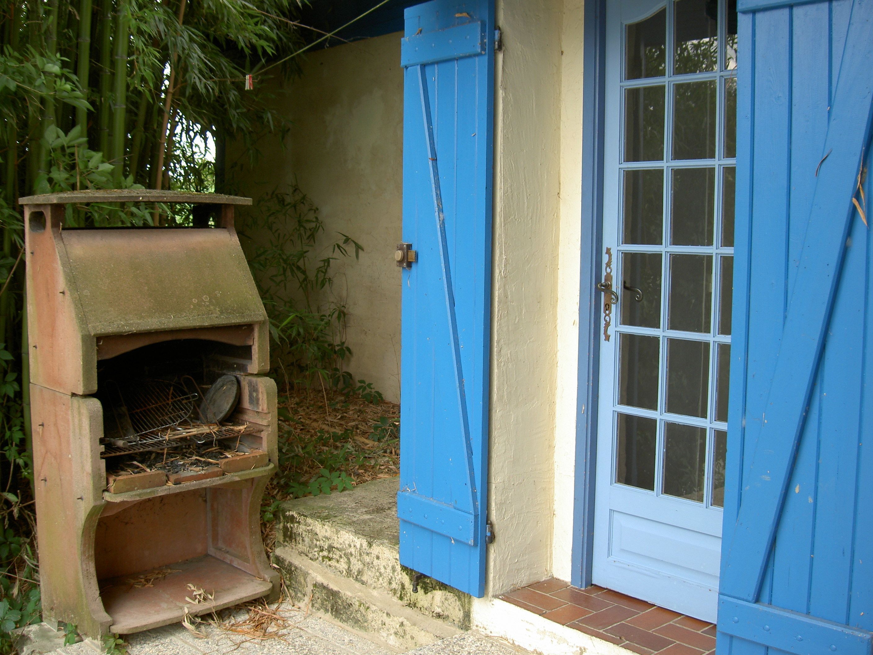 Un barbecue d’extérieur rustique, doté d’une porte et d’une fenêtre bleues, et entouré de verdure.