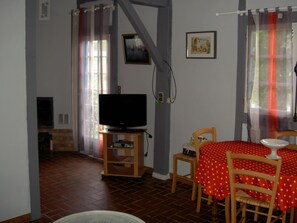 Een kamer met een televisie op een houten standaard, een eettafel met een tafelkleed met rode stippen, en een vloer van baksteen.