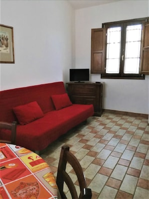 Quarto com sofá vermelho, televisão sobre um móvel de madeira, janela com persianas de madeira e chão em azulejos.