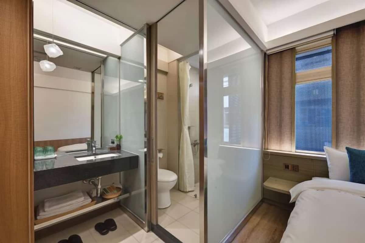 A modern hotel room with a glass partition, a bathroom with a toilet and shower, a bed with white bedding, and a window with curtains.