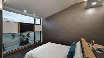 Ein modernes Schlafzimmer mit einem großen Bett, einem Nachttisch aus Holz, einem an der Wand montierten Fernseher und Blick auf die Stadt durch ein großes Fenster.