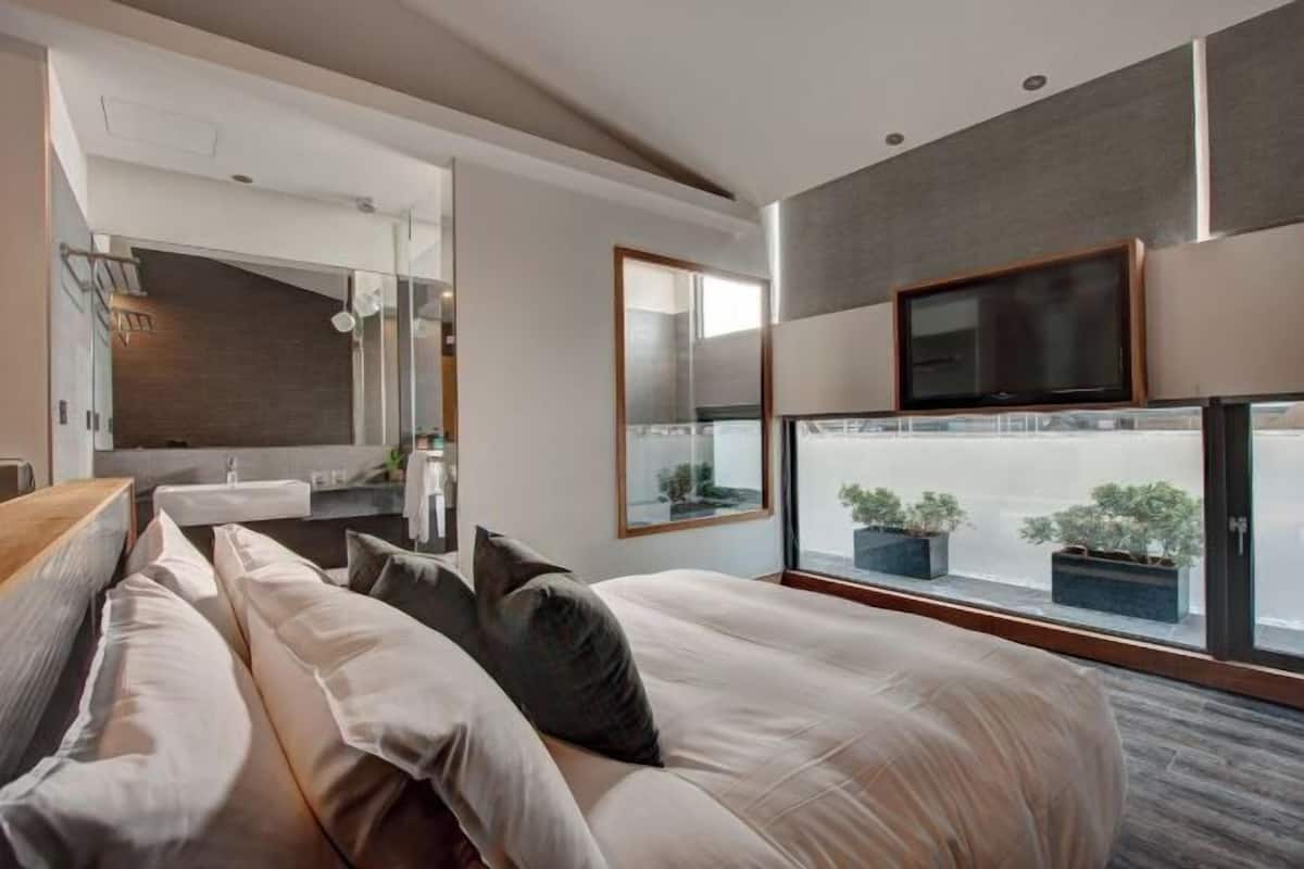 A modern hotel room with a large bed, a TV, and a view of the city through large windows.