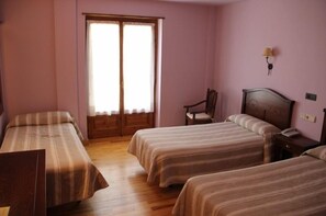 Una habitación con dos camas, piso de madera, ventana con cortinas, una silla y una lámpara de pared.