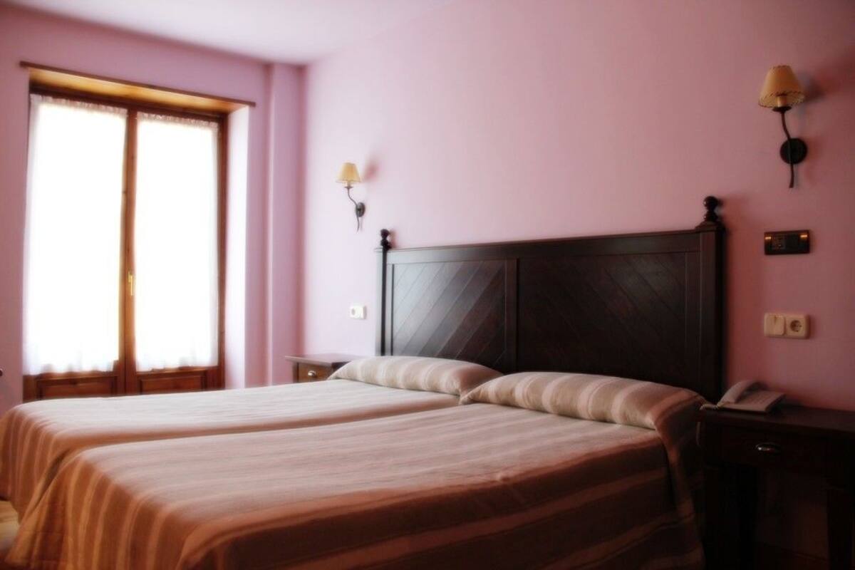 A room with two beds, a window with curtains, a bedside table, and a wall-mounted lamp.