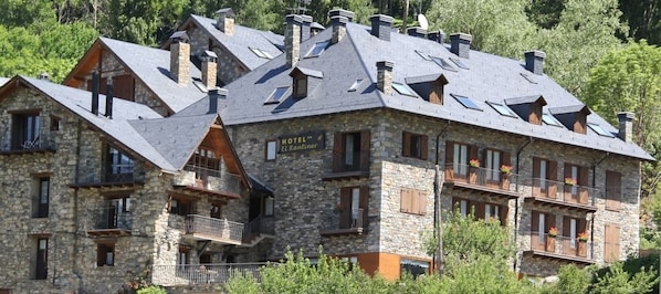 Un edificio de piedra con varios balcones, un letrero que dice “HOTEL EL Rantiner” y un paisaje montañoso de fondo.