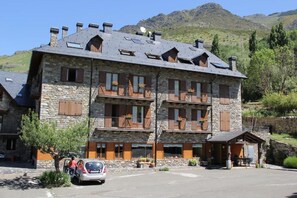 Un edificio de piedra con varios balcones, un coche estacionado al frente y montañas al fondo.