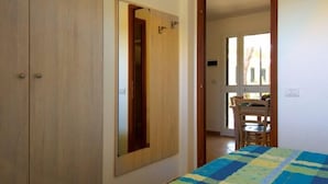 Una habitación con un armario de madera, una cama con colcha azul y amarilla, y un rincón de comedor visible a través de una puerta abierta.