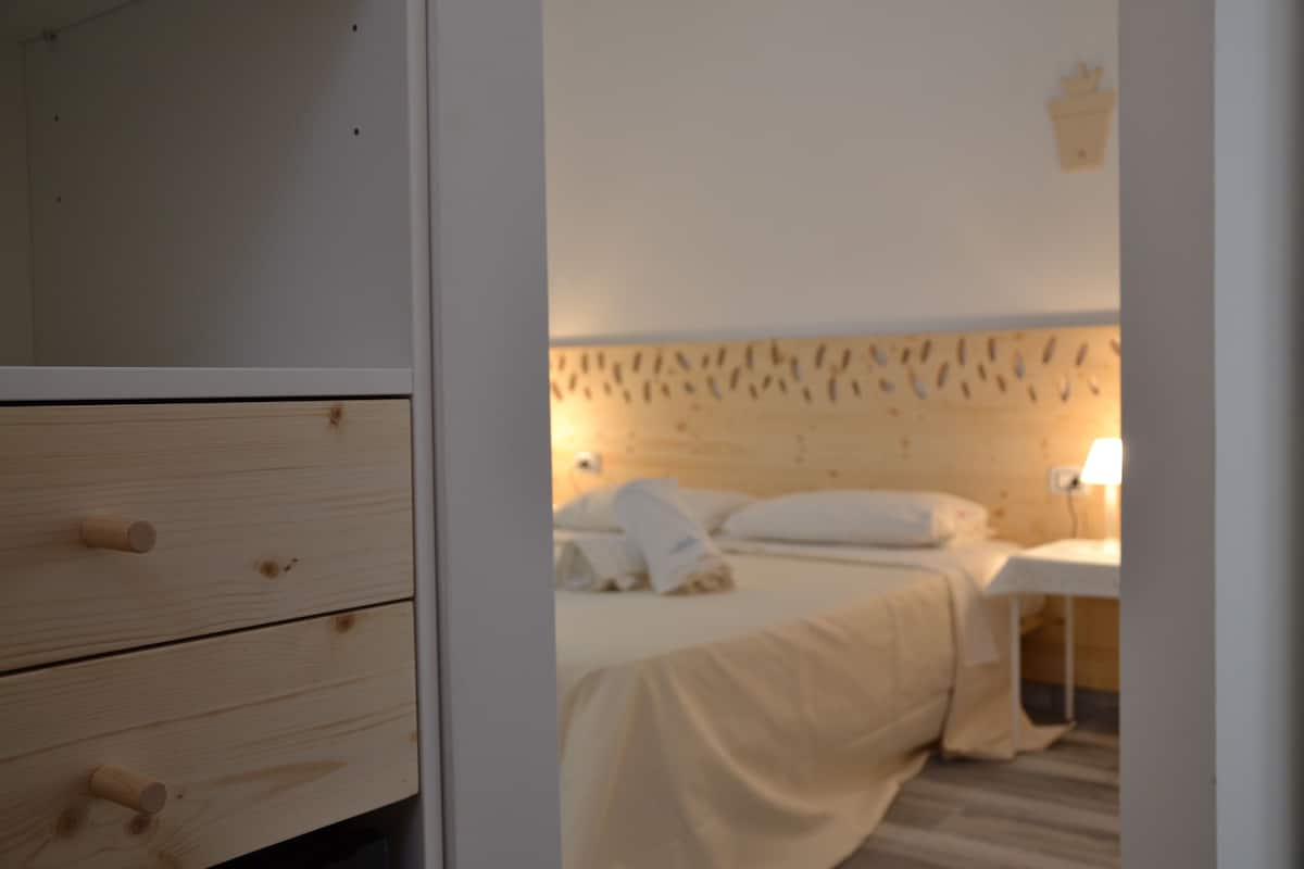 Ein Schlafzimmer mit Holz-Kopfteil, einem Bett mit weißer Bettwäsche, einem Nachttisch mit Lampe und einer Kommode mit Schubladen.
