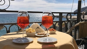 Uma mesa com duas taças de vinho, um recipiente com batatas fritas, um prato de pão e uma pequena tigela de nozes, em uma varanda com vista para o mar.