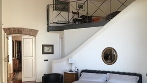 Quarto com cama grande, mesa de cabeceira, cadeira e escada que leva a um andar superior.