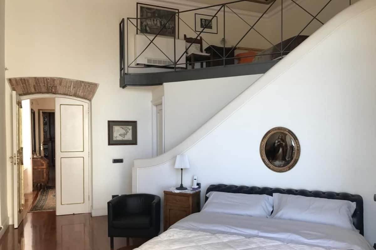 Een slaapkamer met een groot bed, een nachtkastje, een stoel en een trap naar een hoger gelegen verdieping.