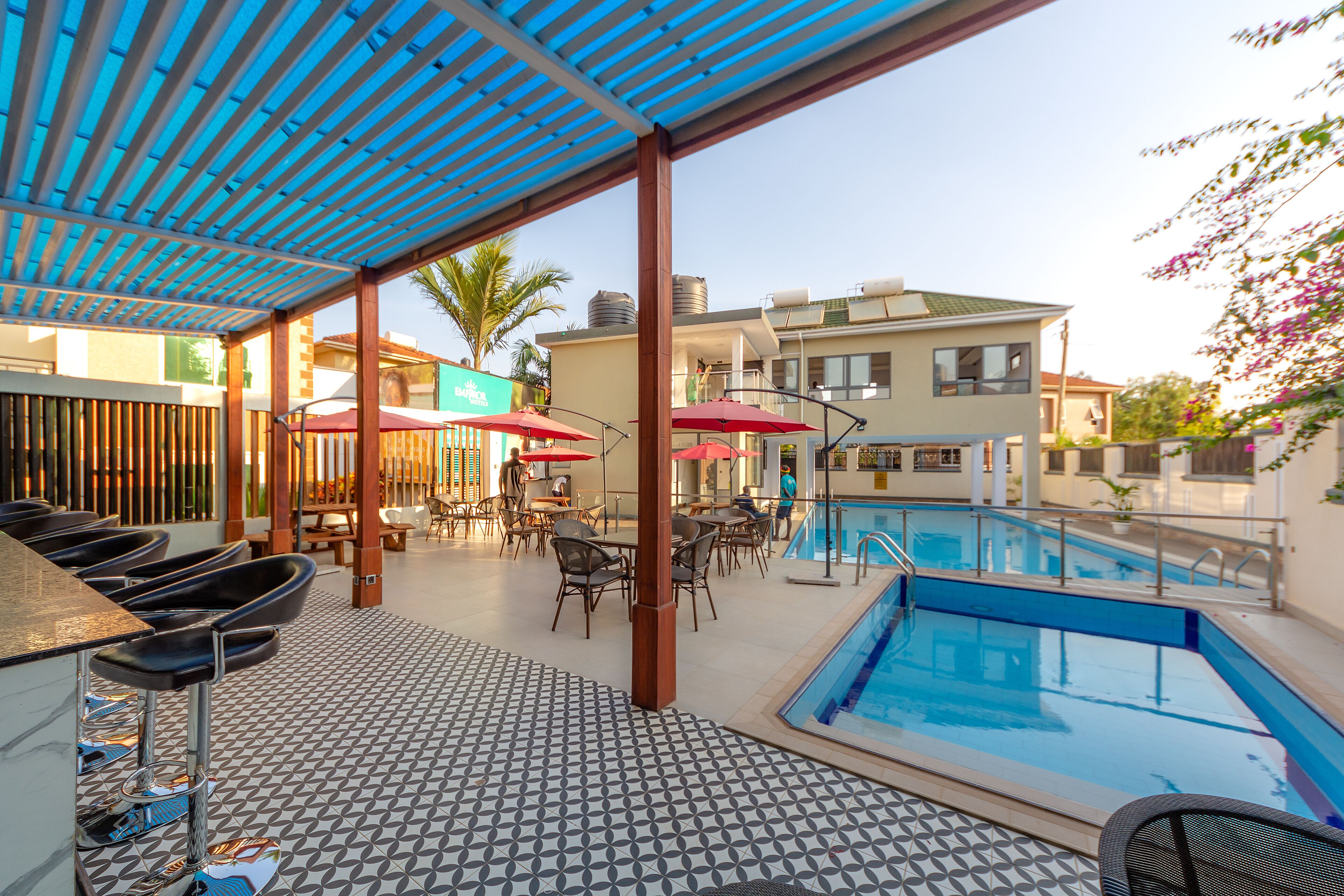A pool area with a tiled floor, a pool, and a covered seating area with chairs and umbrellas.