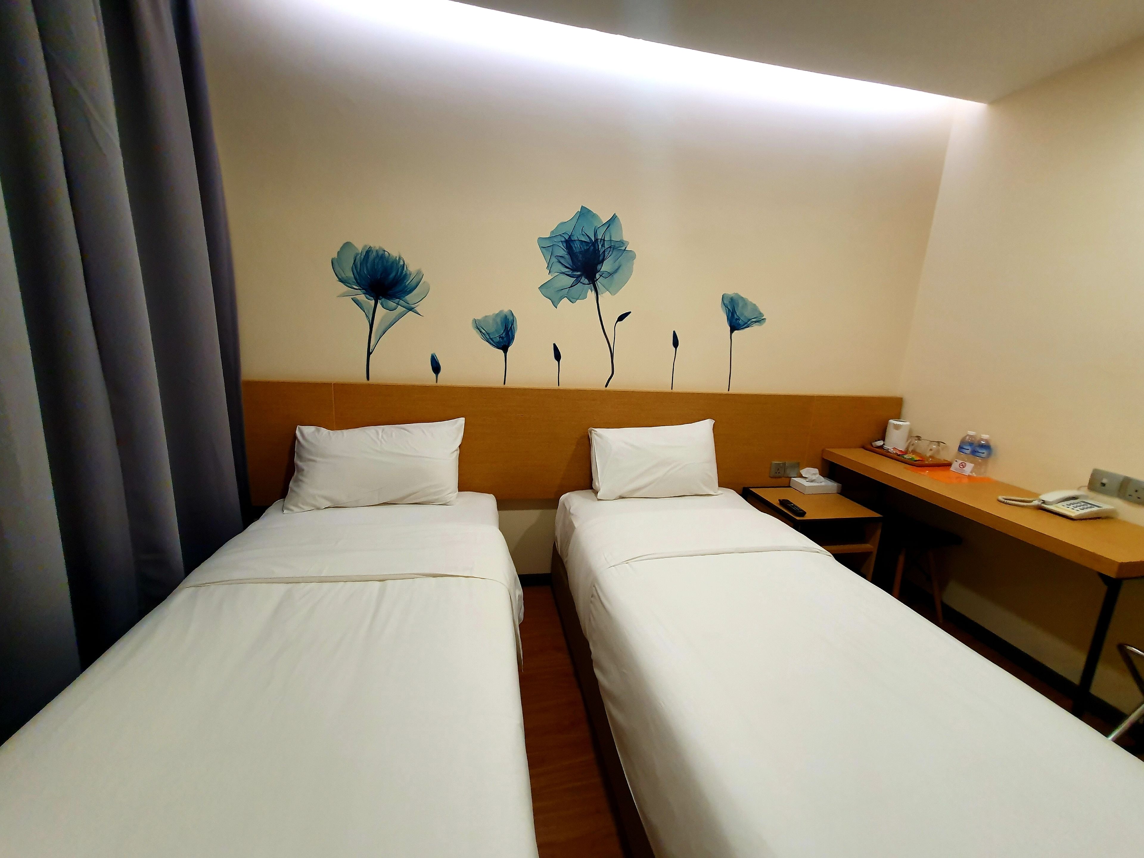 A hotel room with two single beds, a wooden headboard, a desk with a phone, and a wall with floral artwork.