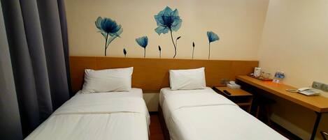 A hotel room with two single beds, a wooden headboard, a desk with a phone, and a wall with floral artwork.