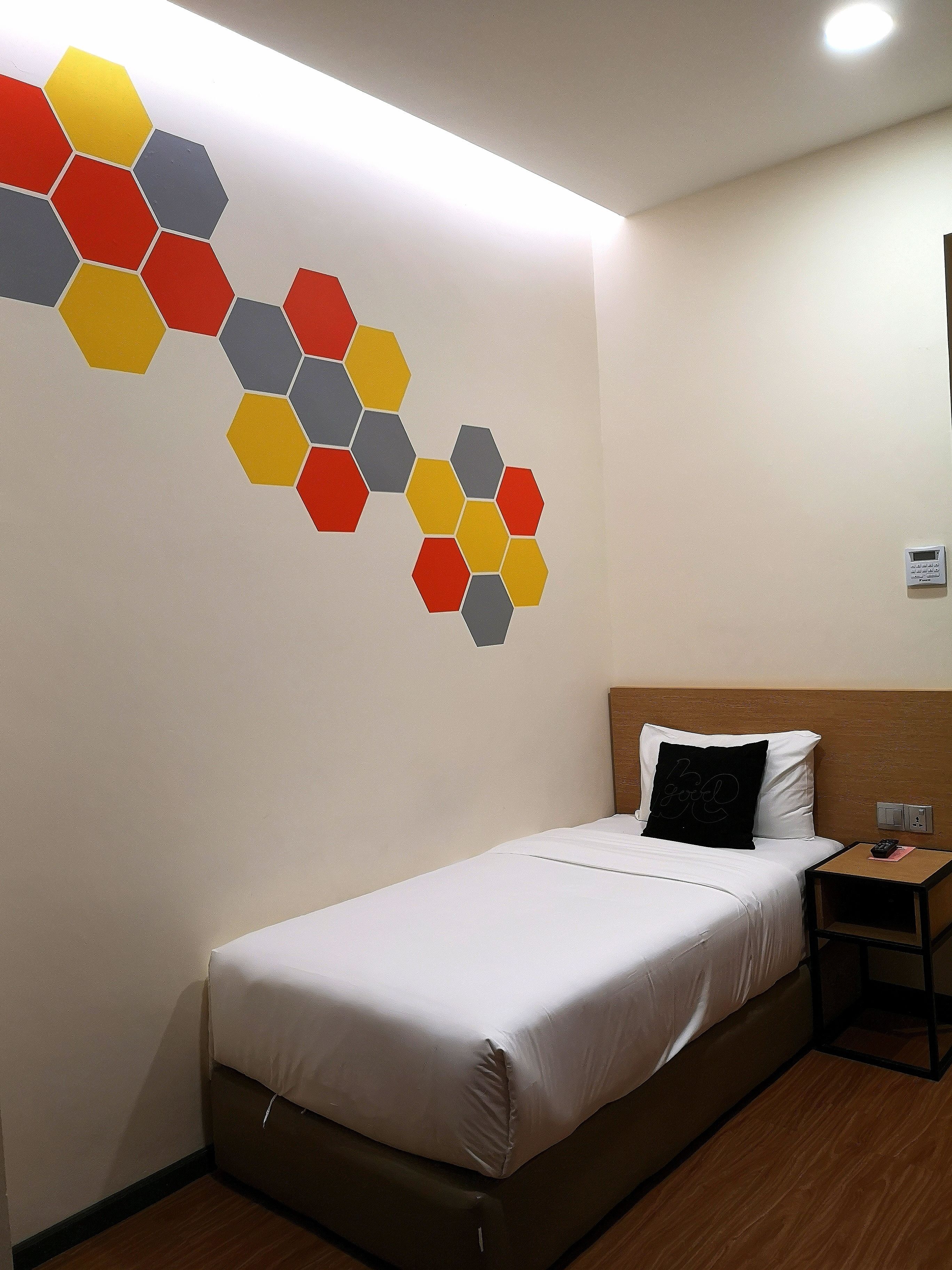 Habitació d'hotel amb un llit, una tauleta de nit, un termòstat fixat a la paret i un dibuix hexagonal decoratiu a la paret.