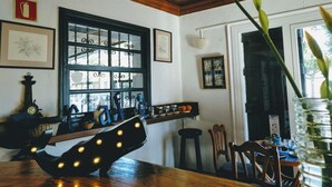 Un'area pranzo con soffitto in legno, un'ampia finestra con inferriate nere e una mensola a parete con diversi oggetti.