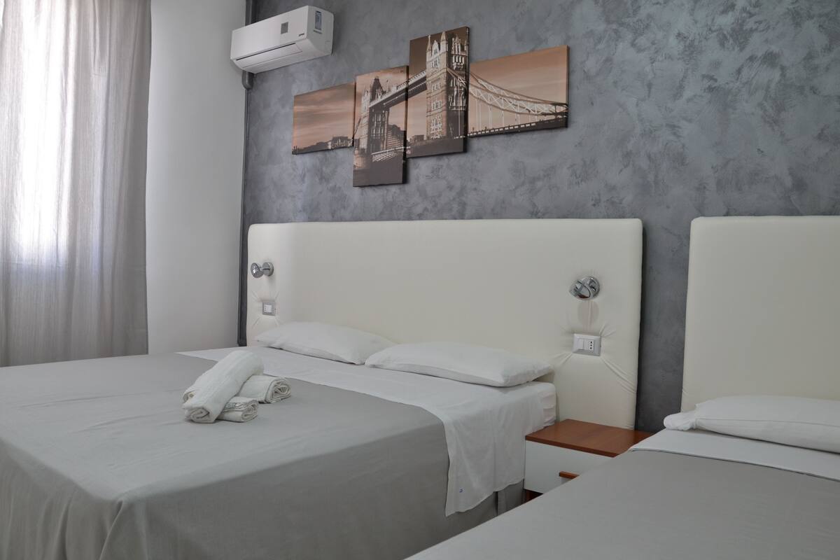 Ein Hotelzimmer mit zwei Betten, einer grauen, strukturierten Wand und einem Gemälde einer Brücke.