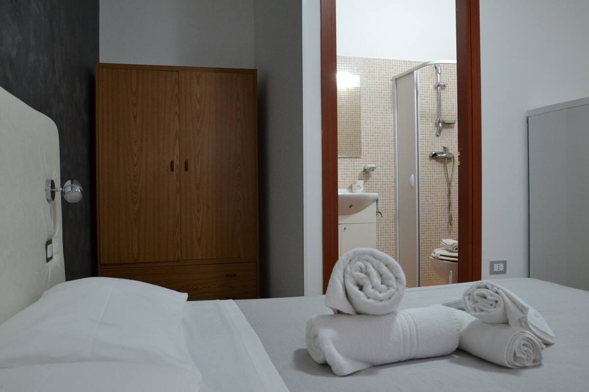 Ein Hotelzimmer mit einem Holzschrank, einem Bett mit weißer Bettwäsche und einem durchgehenden Türrahmen einsehbarer Badezimmer.