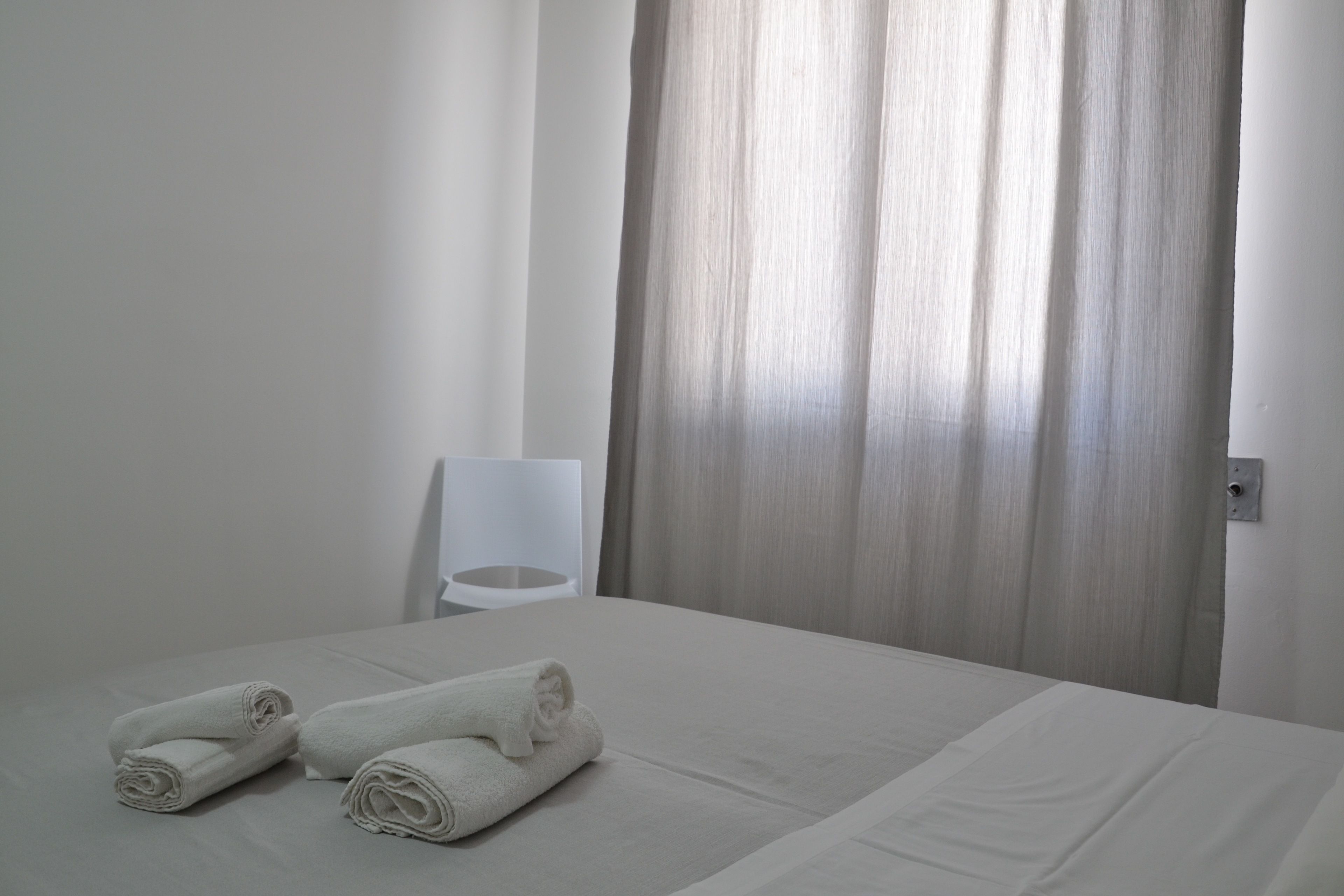 A neatly made bed with rolled white towels, a white chair, and a window with sheer curtains.