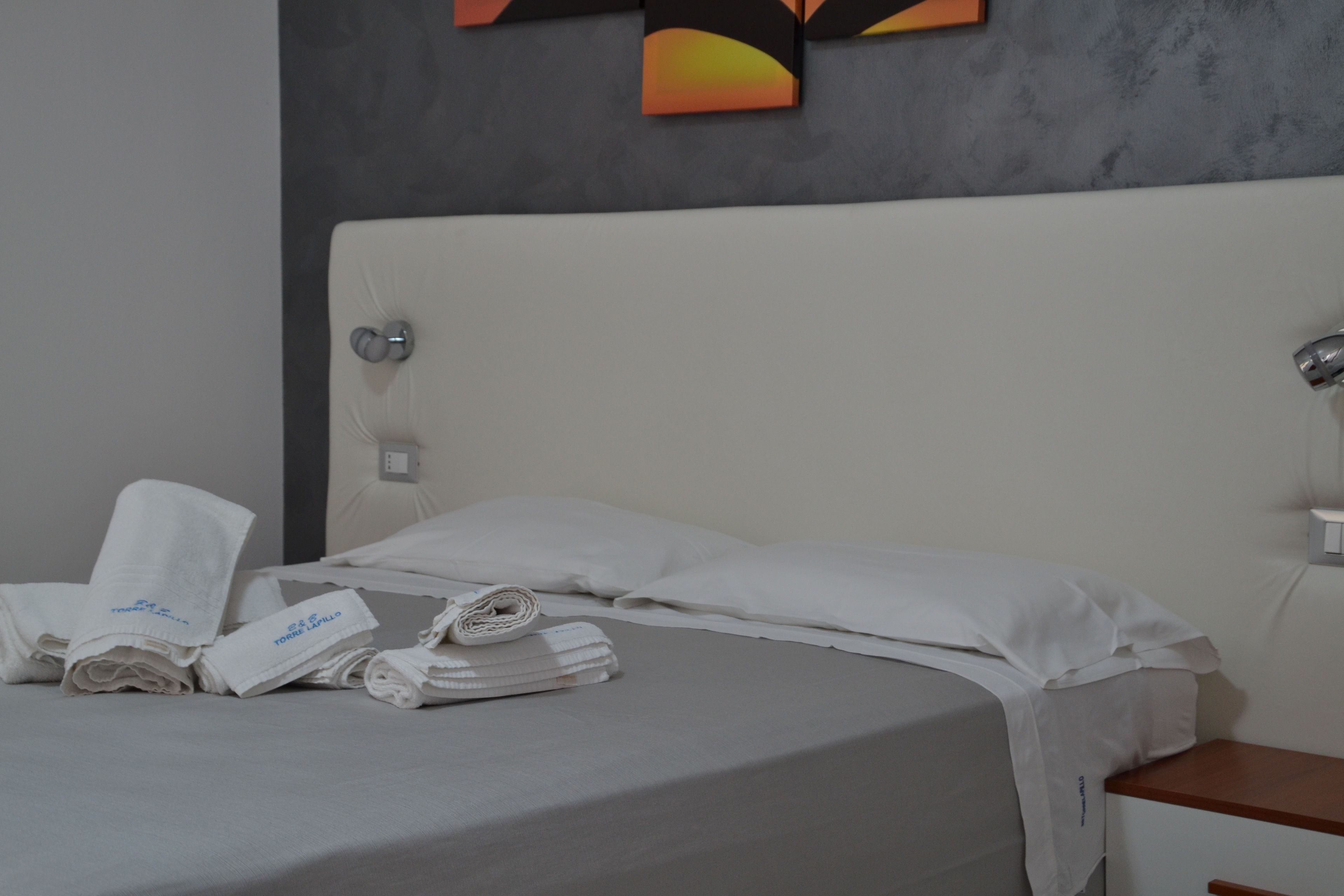 Uma cama bem arrumada com roupa de cama e toalhas brancas, uma cabeceira cinza e uma pequena mesa de cabeceira de madeira com um abajur.