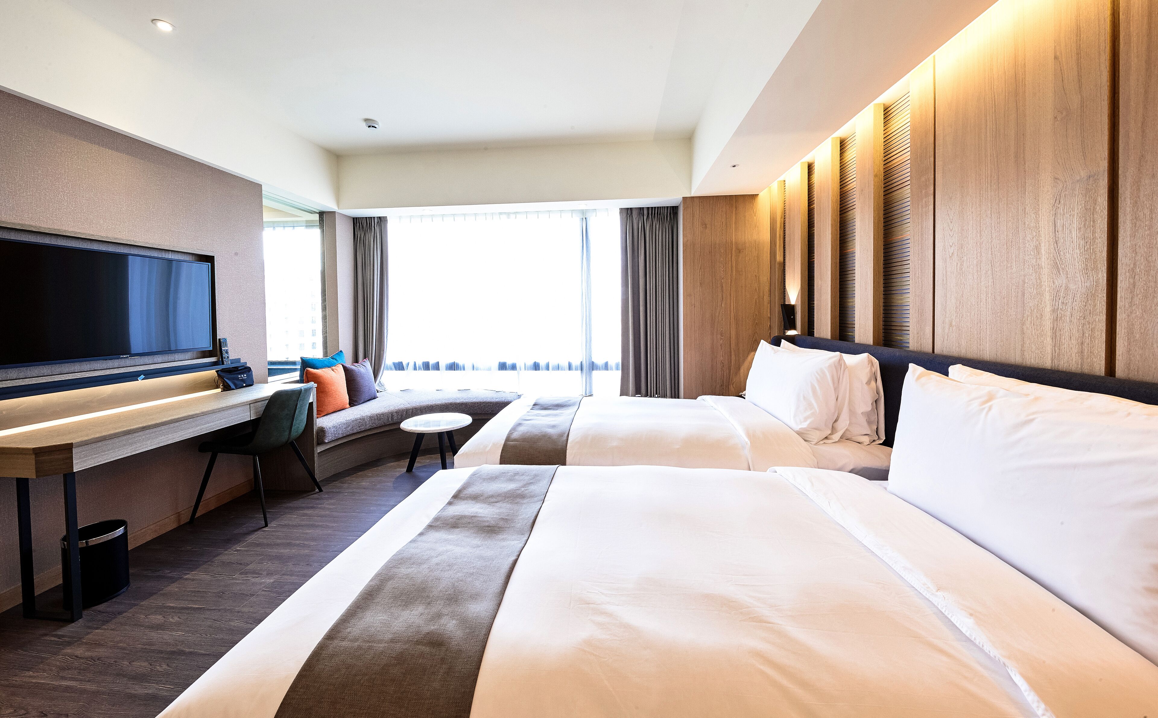 A hotel room with a bed, a desk with a flat-screen TV, a sofa, and a window with curtains.