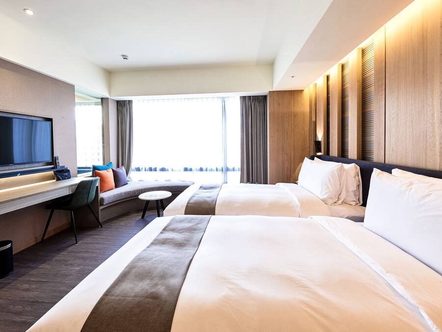 A hotel room with a bed, a desk with a flat-screen TV, a sofa, and a window with curtains.