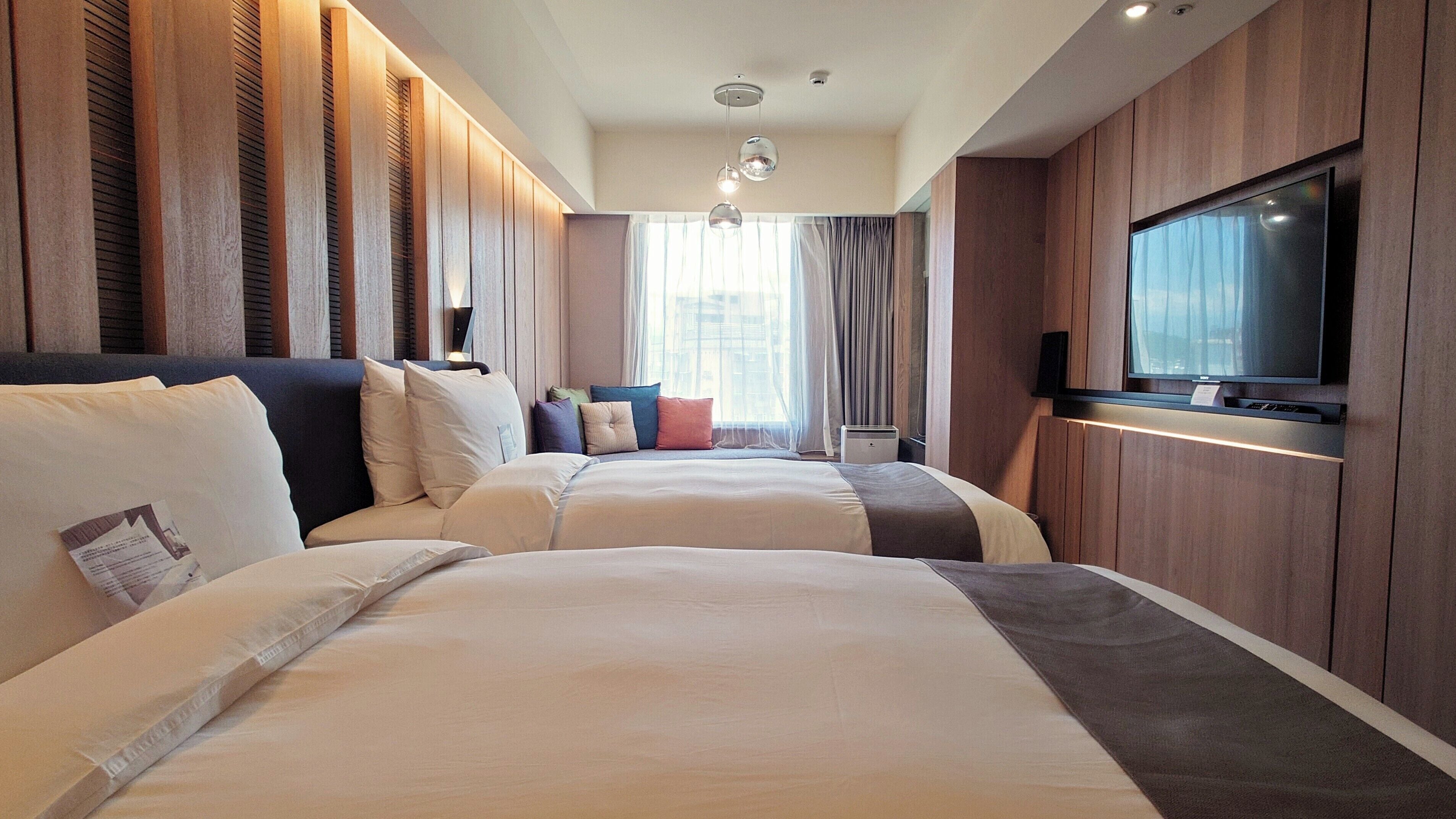 A hotel room with two beds, a flat-screen TV, and a large window with curtains.