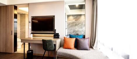 A modern hotel room with a wooden desk, a flat-screen TV, a sofa with colorful cushions, and a large window with curtains.