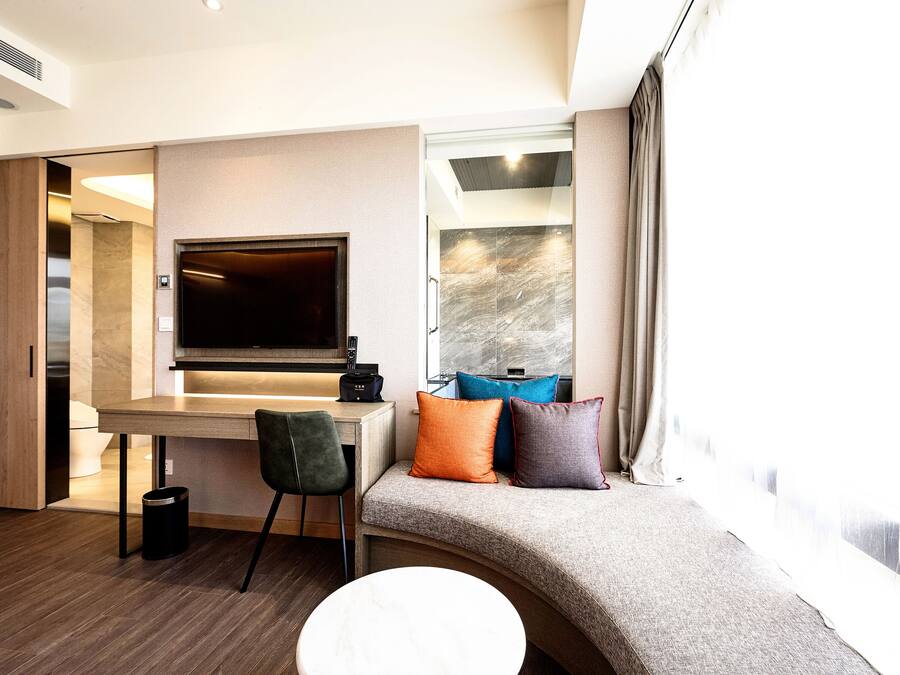 A modern hotel room with a wooden desk, a flat-screen TV, a sofa with colorful cushions, and a large window with curtains.
