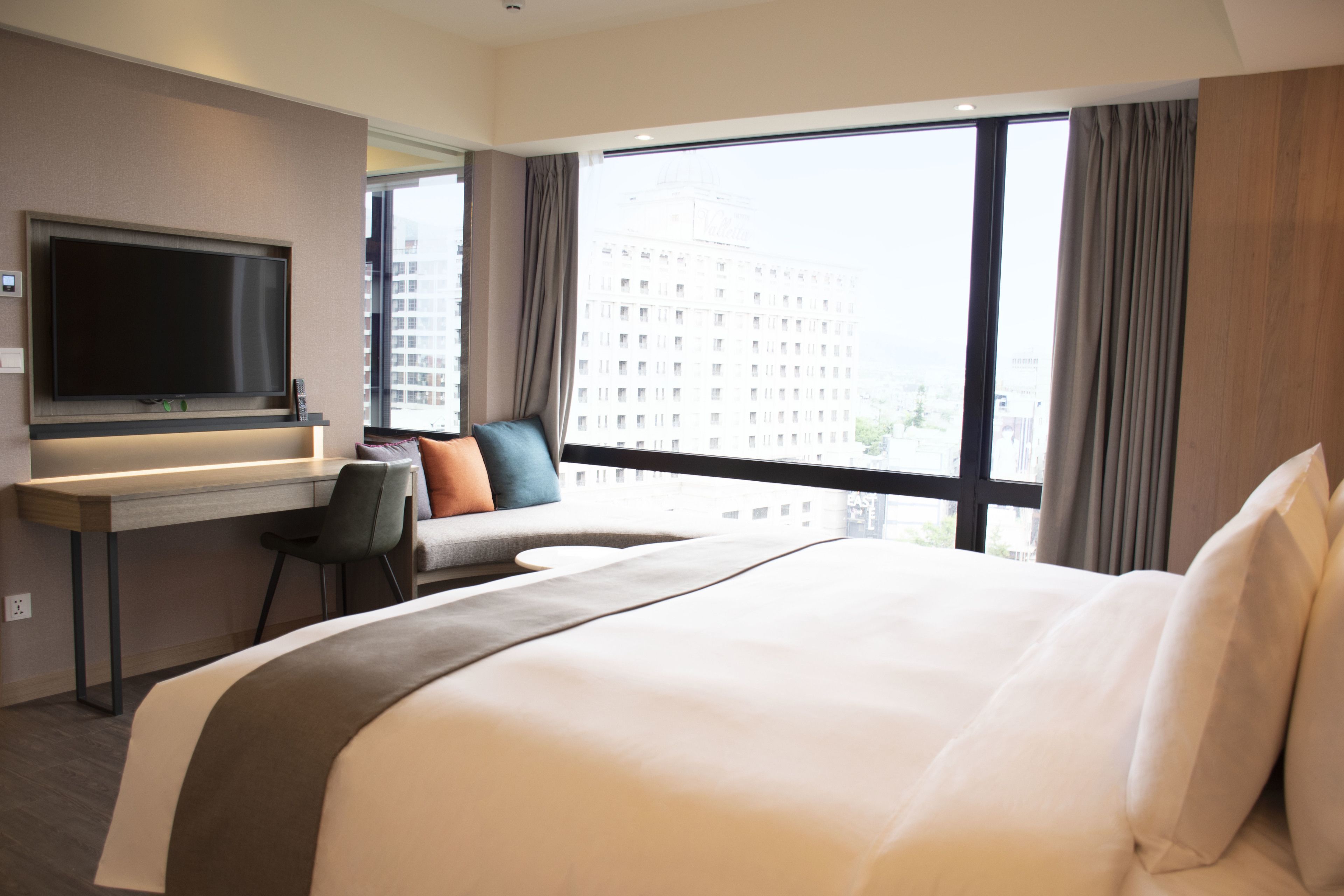 Habitación de hotel con una cama grande, un escritorio, un televisor de pantalla plana, una silla y vistas a un paisaje urbano a través de amplios ventanales.