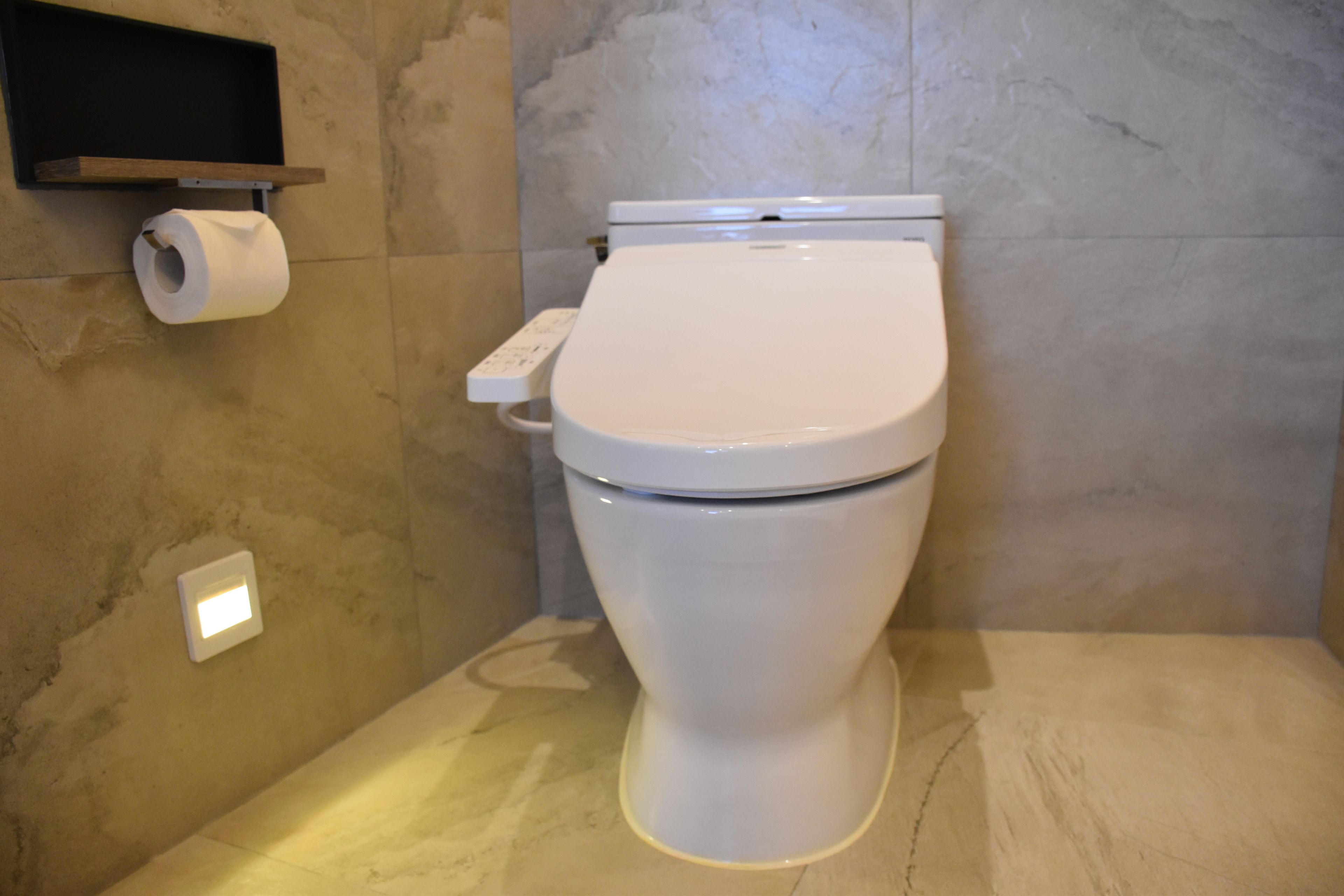 Un baño moderno con inodoro blanco, un dispensador de papel higiénico y un interruptor de luz en la pared.