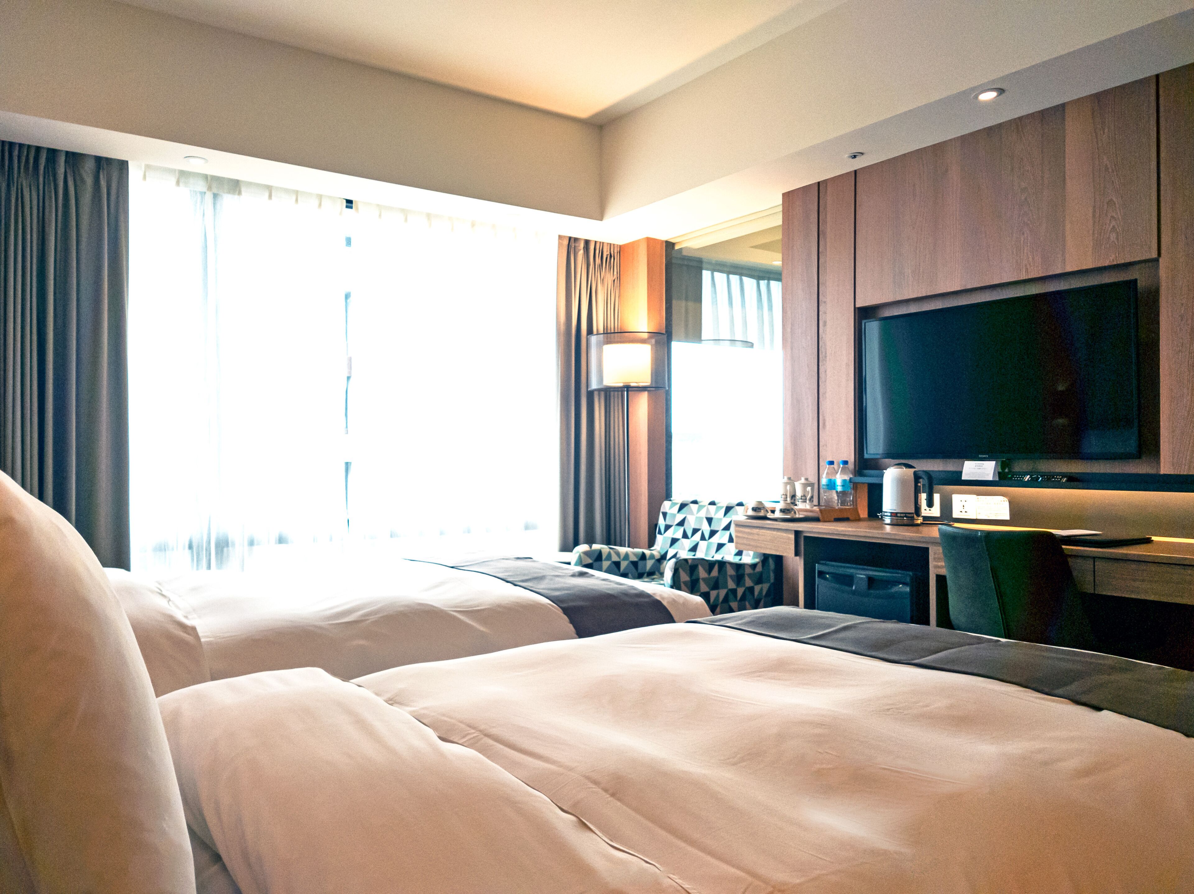 A hotel room with two beds, a desk, a TV, and a window with curtains.