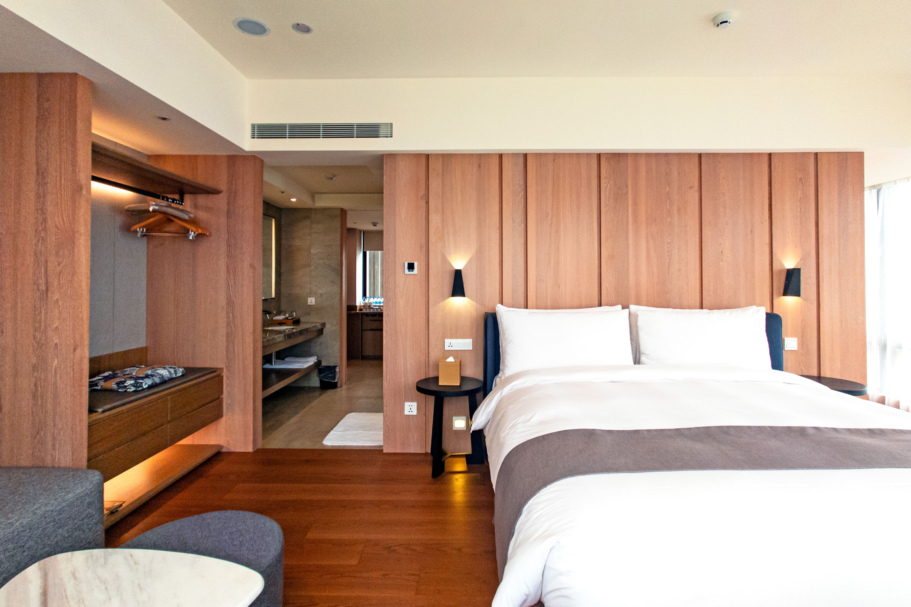 A modern hotel room with a large bed, wooden paneling, a small table with a lamp, and a seating area with a bench and armchair.