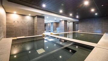 A modern indoor swimming pool with a glass bottom, surrounded by grey tiled walls and a ceiling with recessed lighting.