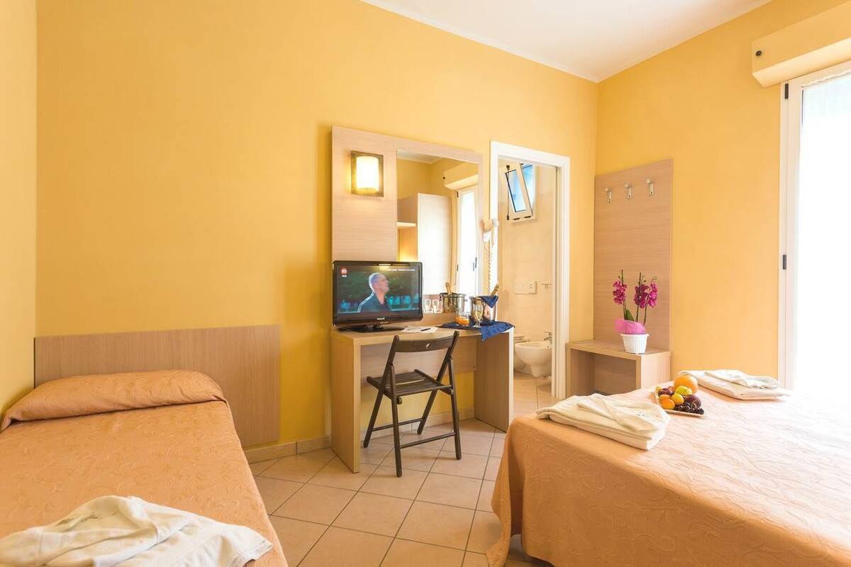 A hotel room with two beds, a desk with a TV, a chair, a small table with toiletries, and a window with a view.