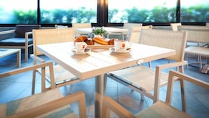 Una cafetería moderna con un diseño limpio y minimalista, que presenta una mesa con pasteles y tazas de café, asientos cómodos y ventanas amplias con vistas a zonas verdes.
