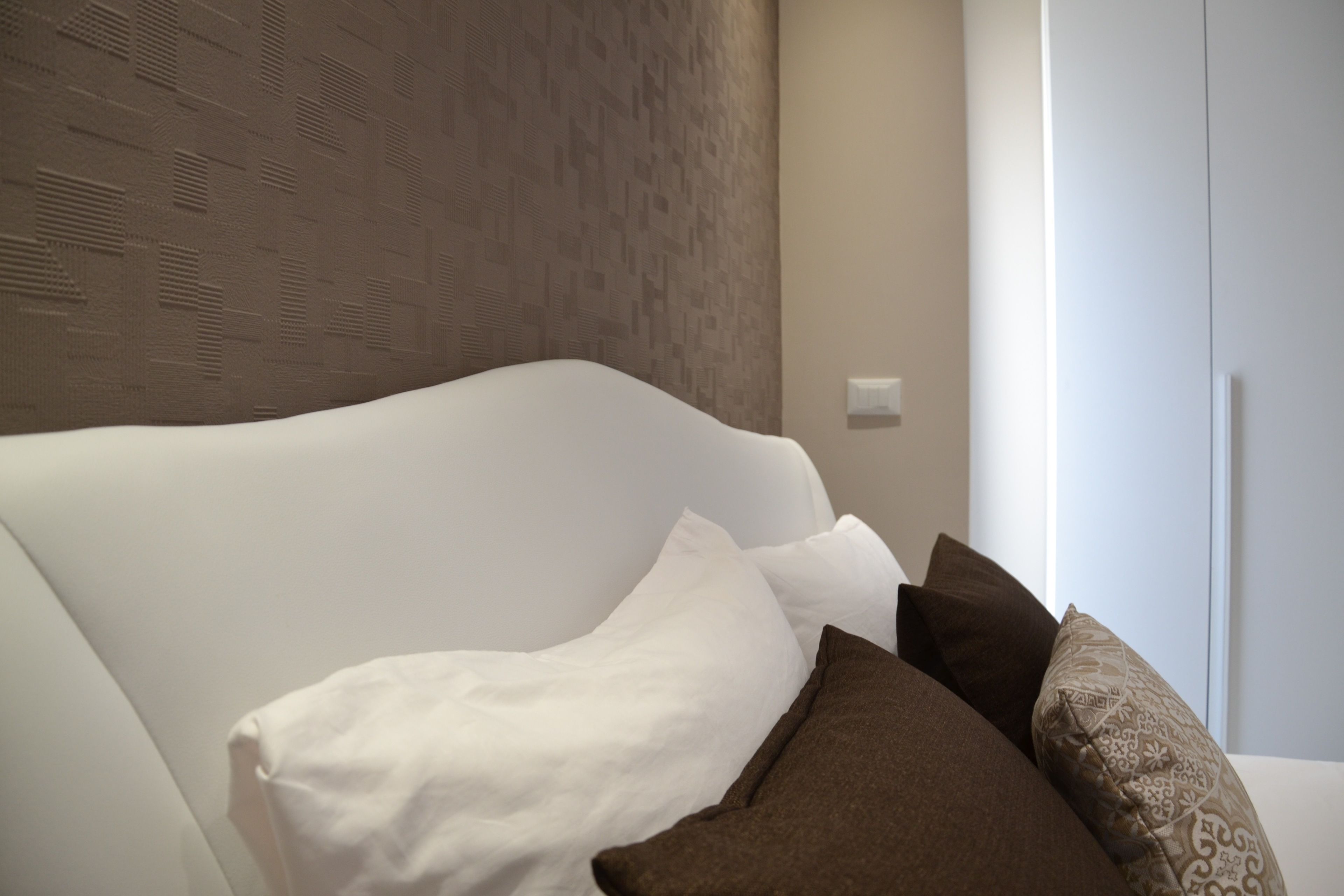 A modern bedroom with a white headboard, a patterned wall, and a white wardrobe.