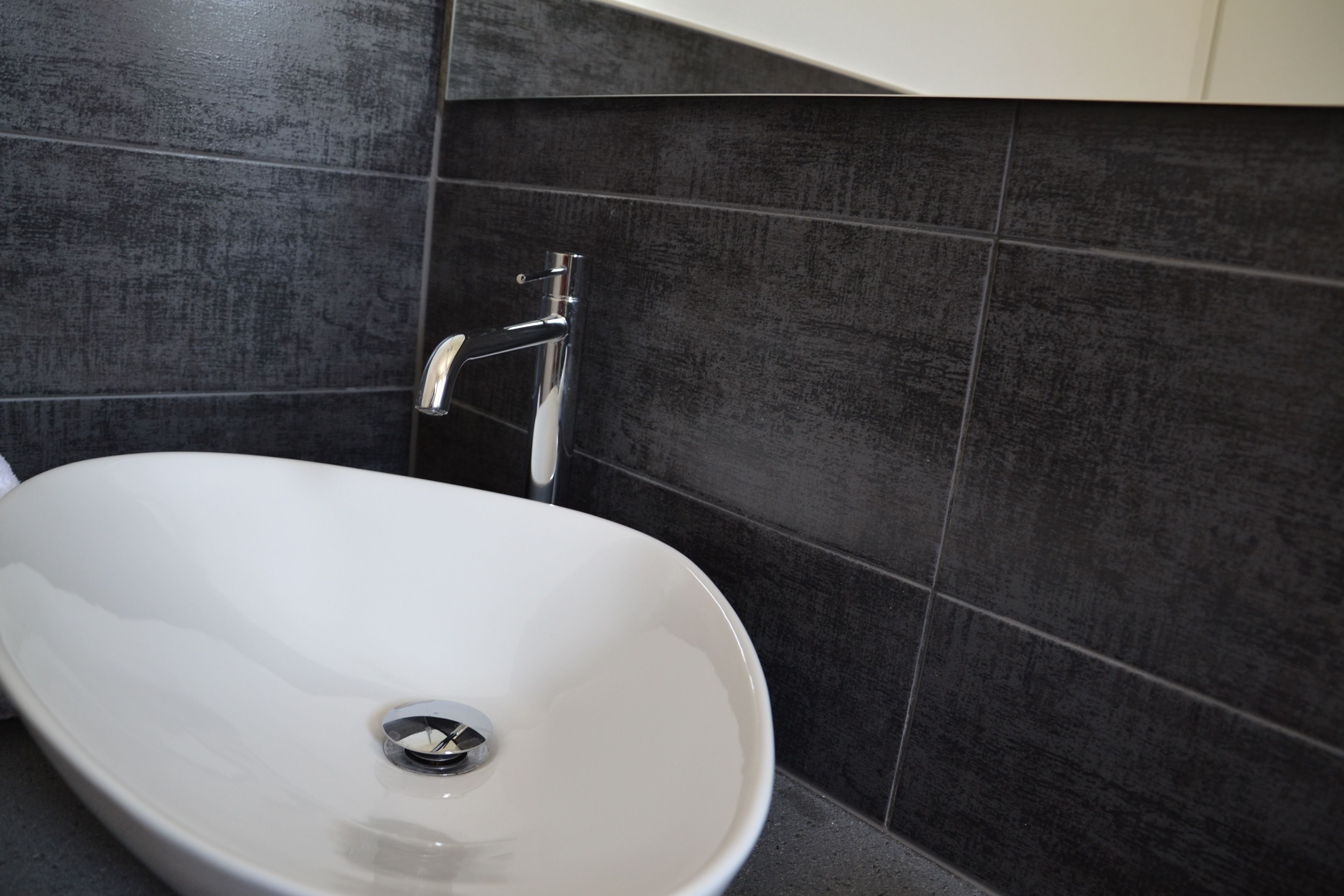 Ein modernes Badezimmer mit einem weißen Waschbecken, dunklen Fliesenwänden und einem Chromarmaturen.