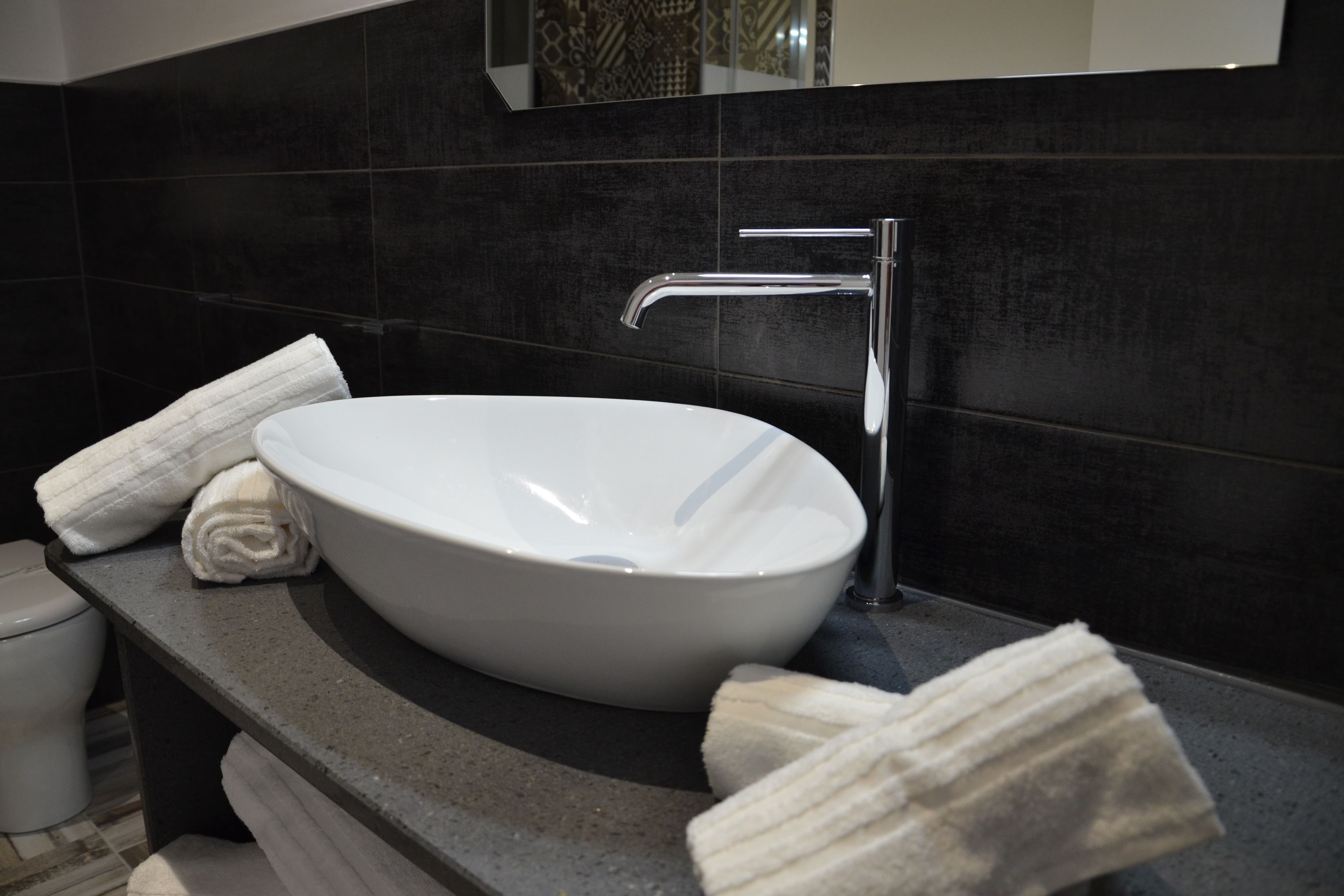 A modern bathroom with a large white vessel sink, a chrome faucet, and dark tiled walls.