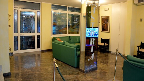 A lobby with a green sofa, a glass partition, a television, and a painting on the wall. - Hotel Astoria (Viareggio)